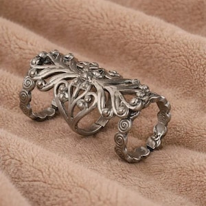Puede incluir: Anillo de dedo plateado y ornamentado con un diseño floral y de volutas. El anillo presenta tres bandas conectadas por una pieza central decorativa. El anillo es de metal y tiene una estética vintage o antigua.