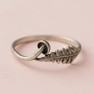 May include: A silver ring with a mushroom and leaf design. The ring has a delicate, nature-inspired aesthetic, with the mushroom cap and leaf details clearly visible. The ring is set against a soft pink background.