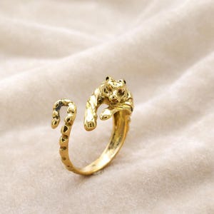May include: A gold-colored ring featuring a detailed tiger design. The ring wraps around the finger with the tiger's head and paws forming the focal point. The ring's band has a textured, dotted pattern.