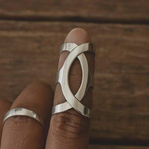 Può includere: Un anello color argento con un design geometrico aperto indossato su un dito. L'anello presenta una forma ovale al centro, con fasce che si intersecano creando un motivo accattivante. Un semplice anello a fascia è visibile anche su un dito adiacente.