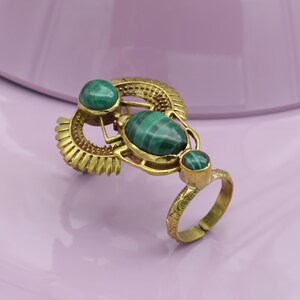 Op de afbeelding: Verstelbare messing ring met drie groene malachietstenen. De ring heeft een sierlijk ontwerp met een ovale centrale steen en twee kleinere ronde stenen. De ring staat tegen een zachte paarse achtergrond.