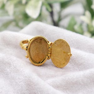 Könnte beinhalten: Goldfarbener Medaillonring mit einem ovalen Fach, das sich öffnet. Der Ring liegt auf einer weißen Oberfläche. Das Medaillon ist geöffnet und gibt den Innenraum frei.