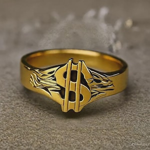 May include: A gold-coloured ring with a dollar sign motif. The ring has a broad band, and the dollar sign is the central design element. The ring is likely made of a gold-toned metal and is set against a neutral background.