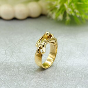 May include: A gold-colored ring with a unique sculptural design. The ring features an abstract figure embracing another, with intricate details and a polished finish. The ring's band is smooth and wide, showcasing the detailed artwork.