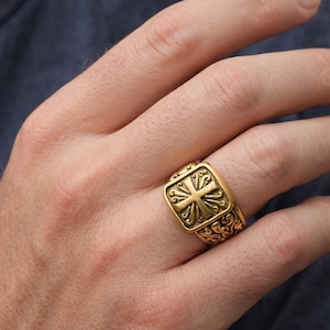May include: A gold-colored ring with a square face featuring a cross design. The ring has an ornate, scrolled pattern on the band and around the face. The ring is on a person's finger.