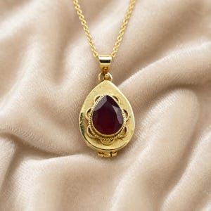 May include: A gold-toned locket pendant on a matching delicate chain. The locket is teardrop-shaped with a central, faceted, deep red stone surrounded by a decorative, twisted metal border. The piece features a hinged design at the bottom.