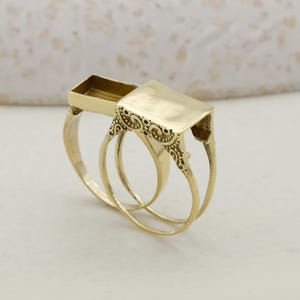Könnte beinhalten: Ein goldfarbener Ring mit rechteckiger Oberseite und aufwendigen Filigranarbeiten. Der Ring hat ein Doppelband-Design und ein kleines, offenes rechteckiges Fach. Der Ring ist wahrscheinlich aus Messing oder einem ähnlichen Metall gefertigt.