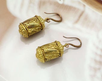 May include: A pair of gold-colored earrings with a cylindrical design. The earrings feature intricate leaf and scroll patterns, with a hook closure. The earrings have a vintage or antique aesthetic.