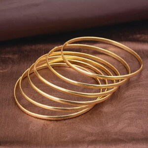 Può includere: Una collezione di braccialetti color oro, impilati e disposti su una superficie marrone. I braccialetti sono sottili e circolari, riflettendo la luce. L'immagine mostra i gioielli su uno sfondo complementare, evidenziando la lucentezza metallica dei braccialetti.