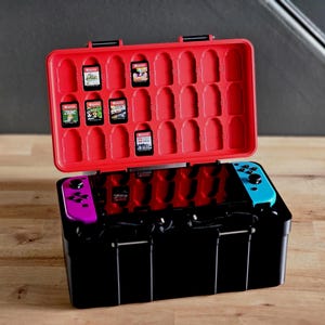 Pode incluir: Uma caixa preta e vermelha para console de jogos com interior vermelho, contendo cartuchos de jogos e um console. A caixa está aberta, revelando o console com controles roxos e azuis. A caixa está em uma superfície de madeira.