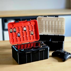 Pode incluir: Estojo de transporte preto e vermelho para console de jogos com um controle preto. A caixa está aberta, revelando cartuchos de jogos. Outro estojo é visível, junto com um console de jogos.