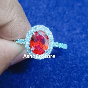 May include: A silver ring featuring a large, oval-cut, vibrant red gemstone, encircled by a halo of smaller, clear stones. The ring band is also set with small, clear stones. The text "AshnCostore" is visible.