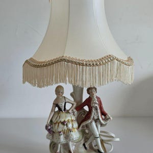 Op de afbeelding: Een tafellamp met een porseleinen voet met twee figuren in historische kleding, een persoon in een bloemenrok en een persoon in een rode jas. De lamp heeft een crèmekleurige stoffen kap met een decoratieve goudkleurige rand en franjes.
