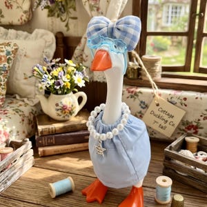 May include: A white goose figurine wearing a light blue gingham bow, light blue glasses, and a light blue dress with a pearl necklace featuring a star charm. A tag attached to the goose reads Cottage Duckling Co. 7" tall.