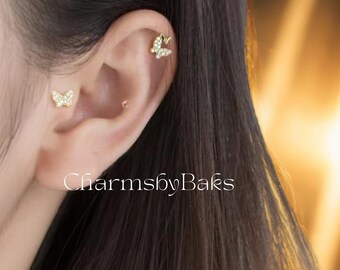 May include: Gold-toned butterfly earrings, one stud and one helix, encrusted with small, clear crystals. The earrings are on an ear, with the text "CharmsbyBaks" in white at the bottom.