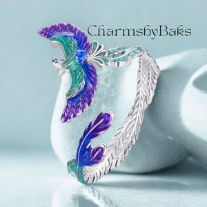 May include: A silver-toned bracelet with a phoenix design. The phoenix is detailed with blue, teal, and purple enamel. The bracelet features a feather design on the band and the text "CharmsbyBaks".