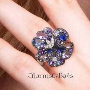 May include: A flower-shaped ring with a silver band. The ring features a cluster of purple, blue, and clear gemstones. The text "CharmsbyBaks" is at the bottom of the image.