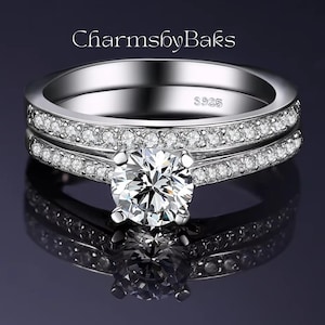 Puede incluir: Un juego de anillos de plata con una piedra central grande, clara y de talla redonda y piedras más pequeñas y claras a lo largo de las bandas. El anillo está marcado con "S925". El texto "CharmsbyBaks" está en la parte superior.