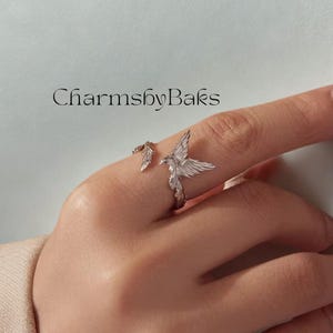 May include: A silver ring with a bird design, wings spread wide. The ring wraps around the finger, with a feather detail. The text "CharmsbyBaks" is visible in the image.