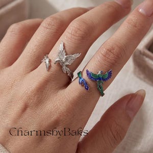 Peut inclure: Deux bagues ouvertes portées sur les doigts. Une bague est de couleur argentée avec un motif d'oiseau. L'autre bague présente un oiseau avec des détails en émail bleu et vert sur ses ailes et sa queue. Le texte CharmsbyBaks est visible en bas à gauche.