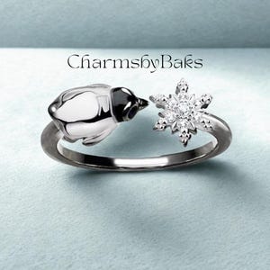 Puede incluir: Un anillo plateado con un diseño de pingüino y copo de nieve. El pingüino tiene la cabeza negra y el cuerpo blanco. El copo de nieve está adornado con pequeñas piedras brillantes. El anillo está sobre un fondo azul claro. El texto en la imagen dice "CharmsbyBaks".