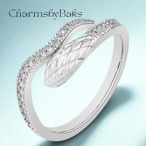 Puede incluir: Anillo plateado con forma de serpiente. La cabeza de la serpiente tiene textura y el cuerpo se curva alrededor del dedo. El anillo está adornado con pequeñas piedras preciosas transparentes. El texto "CharmsbyBaks" está en la esquina superior izquierda.