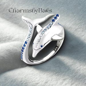 Puede incluir: Un anillo de plata con diseño de delfín y gemas azules y transparentes. El anillo presenta un diseño de delfín, con la cola y el cuerpo que envuelven el dedo. El anillo está marcado con "925" y el texto "CharmsbyBaks".
