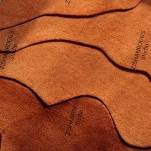 May include: An abstract rug with a wavy design in shades of orange and brown. The rug has a textured surface and the text "ZISHANRUGGS Studio" is printed on the side.