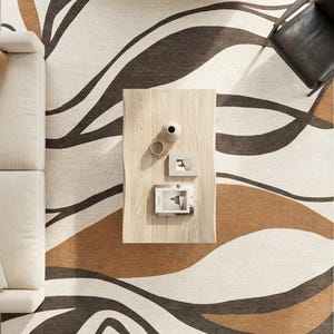 Könnte beinhalten: Draufsicht auf einen Wohnzimmerteppich mit modernem Design. Der Teppich zeigt fließende, abstrakte Muster in Braun-, Beige- und Weißtönen. Ein helles Sofa, ein Holztisch und ein schwarzer Stuhl sind ebenfalls zu sehen.