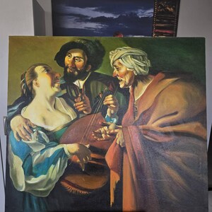 May include: Oil painting depicting three figures, a man playing a lute, a woman, and an older person. The figures are dressed in period clothing, with the woman in a blue and white dress. The painting has a dark background and is mounted on a wooden easel.