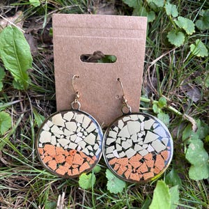 May include: A pair of round dangle earrings featuring a mosaic design of cream and terracotta-colored fragments encased in a clear resin within a gold-toned metal frame, suspended from gold-toned ear wires on a brown rectangular display card.