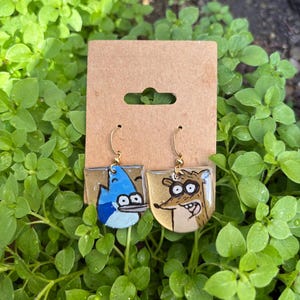 May include: A pair of dangle earrings featuring hand-painted illustrations of two cartoon characters. One earring depicts a blue jay with a white face, and the other shows a brown raccoon with large eyes. Both are attached to gold-toned metal hooks.