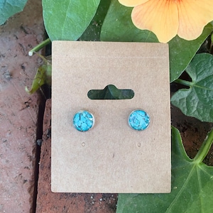 May include: A pair of round stud earrings with a silver-colored setting. Each earring features a vibrant turquoise stone with a mosaic pattern. The earrings are displayed on a brown cardboard earring card.