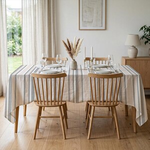 May include: A dining table set for four with a striped tablecloth in beige, grey, and white. The table is set with plates, glasses, and silverware. Wooden chairs surround the table, and a vase of dried flowers sits in the centre.