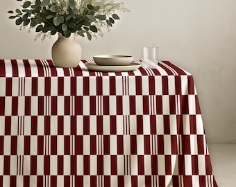 May include: A rectangular tablecloth featuring a repeating geometric pattern of deep red vertical rectangles and white stripes. On the table sits a beige ceramic vase filled with green eucalyptus branches, a shallow beige bowl, and a clear glass tumbler.