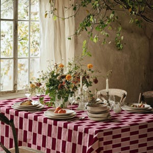 Può includere: Un tavolo da pranzo apparecchiato per un pasto, con una tovaglia a scacchi rossi e rosa. Il tavolo è apparecchiato con piatti bianchi, bicchieri e un centrotavola floreale con fiori arancioni e bianchi. Sono visibili due candele accese.