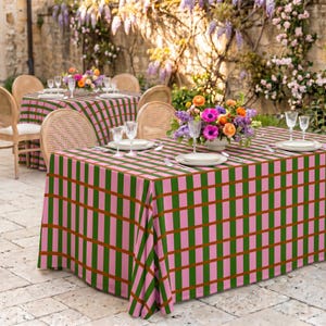 May include: A rectangular tablecloth featuring a grid pattern of pink and green vertical and horizontal stripes with brown accent lines. The tablecloth is draped over a table set with white plates, clear glassware, and a floral centerpiece.