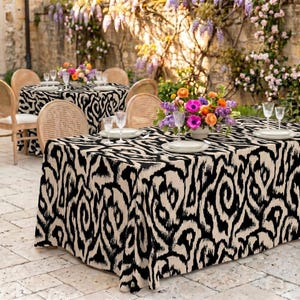 May include: A rectangular tablecloth featuring a bold, black and beige abstract swirl pattern. The fabric drapes over a table set with white plates, clear glassware, and a vibrant floral centerpiece, creating a cohesive look for dining.