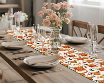 May include: A long rectangular table runner featuring a repeating pattern of orange pumpkins, green leaves, and small yellow berries on a white background. The runner is centered on a wooden table set with white plates, clear glasses, and a glass vase.