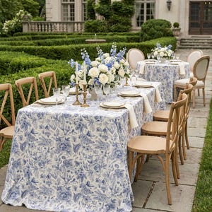May include: A rectangular table covered in a blue and white floral toile tablecloth, set with white plates, clear glasses, and gold candlesticks. A large centerpiece of white roses and blue delphiniums sits in the center, surrounded by wooden chairs.
