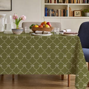 May include: A rectangular tablecloth in a muted olive green color featuring a repeating white lattice pattern with small bow accents at each intersection. The fabric drapes over a dining table set with a glass pitcher, a bowl of fruit, and napkins.