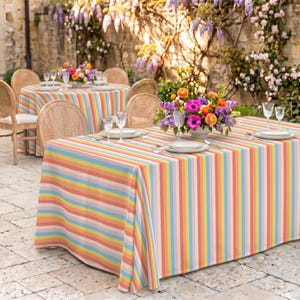 Könnte beinhalten: Ein rechteckiger Tisch mit einer mehrfarbigen, gestreiften Tischdecke, die vertikale Bänder in Pastellrosa, Gelb, Blau und Orange aufweist. Der Tisch ist mit weißen Tellern, klaren Gläsern und einem Blumenarrangement gedeckt.