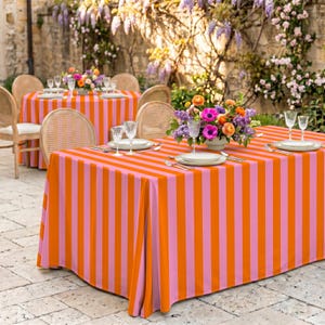 Puede incluir: Una mesa de comedor cubierta con un mantel a rayas naranja y rosa. La mesa está puesta con platos blancos, cubiertos y vasos transparentes. Un arreglo floral está en el centro, y la escena está ambientada al aire libre.