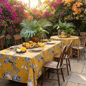 Puede incluir: Una mesa de comedor al aire libre con un mantel amarillo adornado con un estampado tropical en blanco y negro. La mesa está llena de fruta fresca y vajilla. Sillas de madera están dispuestas alrededor de la mesa, sobre un fondo de flores vibrantes y exuberante vegetación.