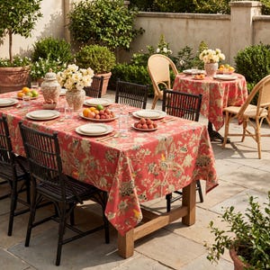 May include: An outdoor dining scene featuring a long wooden table set with plates, glasses, and silverware. The table is covered with a red floral tablecloth. Black chairs surround the table. A smaller round table with a matching tablecloth is also set.