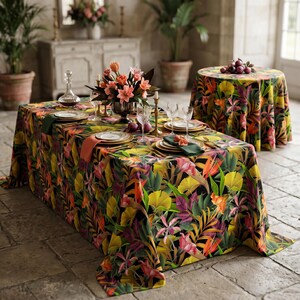 Peut inclure: Une nappe rectangulaire et une nappe ronde avec un motif floral tropical vibrant dans les tons de jaune, orange, rose et vert sur fond noir. La table rectangulaire est dressée avec des assiettes, des verres et une pièce maîtresse florale.