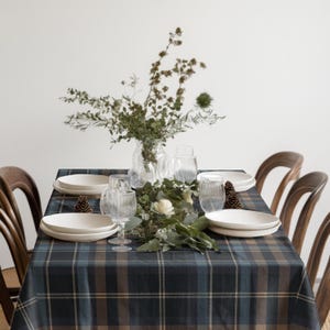 May include: A rectangular dining table covered with a dark blue, brown, and white plaid tablecloth. The table is set with white ceramic plates, clear ribbed glass tumblers, a central floral arrangement in a glass vase, and pinecones.