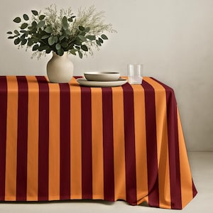 May include: A rectangular tablecloth with vertical stripes in alternating shades of orange and burgundy. A vase with greenery, a bowl, a plate, and a glass of water are on the table.