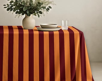 May include: A rectangular tablecloth featuring vertical stripes in alternating shades of deep burgundy and vibrant orange. On top of the tablecloth sits a light-colored ceramic vase filled with green eucalyptus branches, a shallow bowl, and a clear glass.