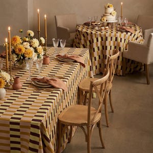 May include: A dining room scene featuring two tables adorned with patterned tablecloths in yellow, brown, and pink. Wooden chairs are arranged around the tables. The tables are set with candles, flowers, and place settings, creating an elegant atmosphere.